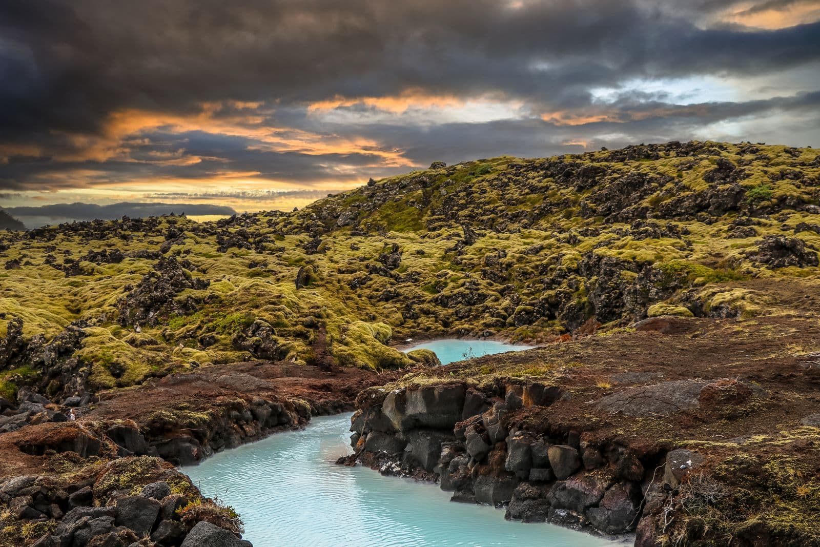 Milky turquoise geothermal water winding through moss-covered lava rock under an orange sunset sky