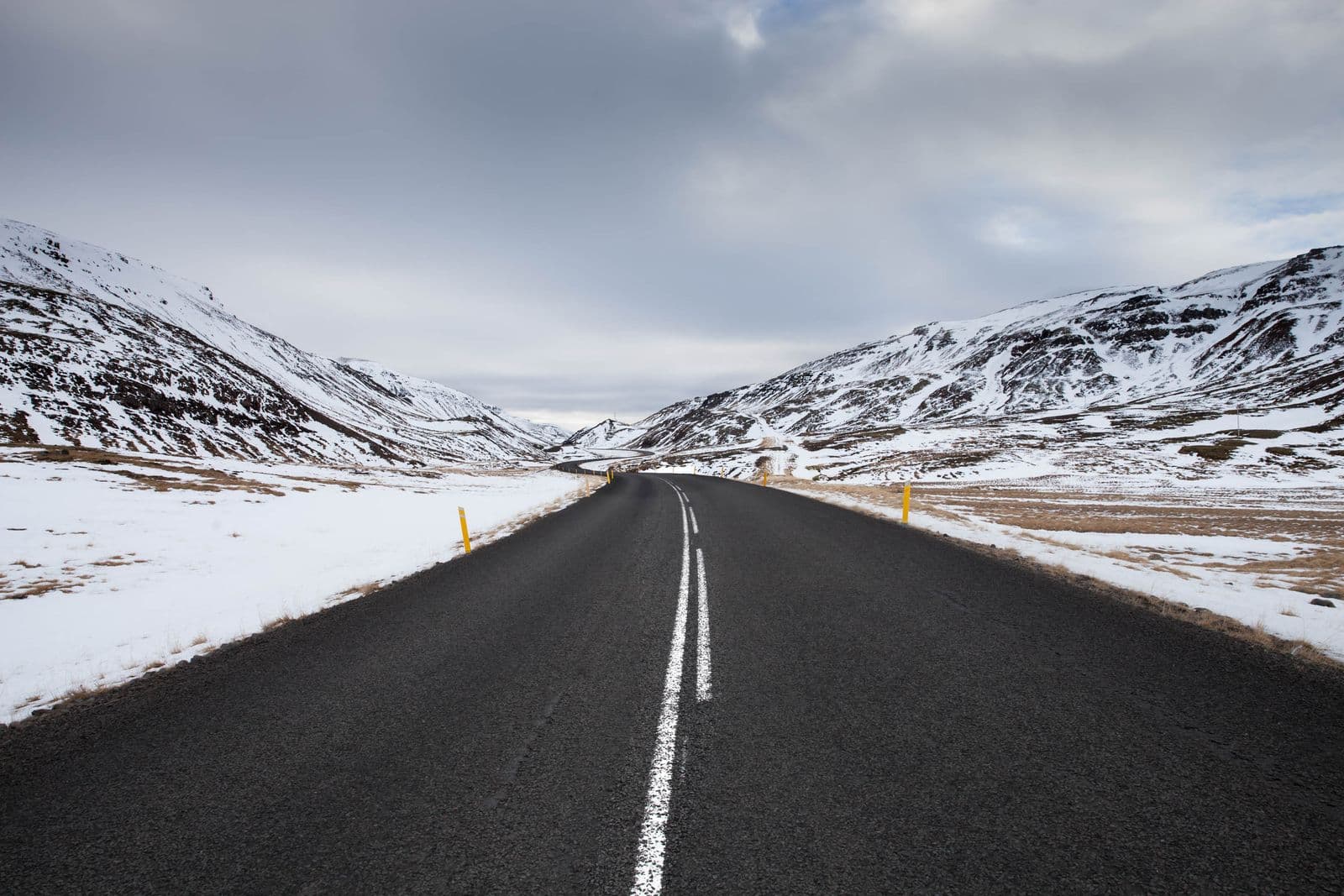 A clear road winding between snow-covered mountains under a cloudy sky in Iceland