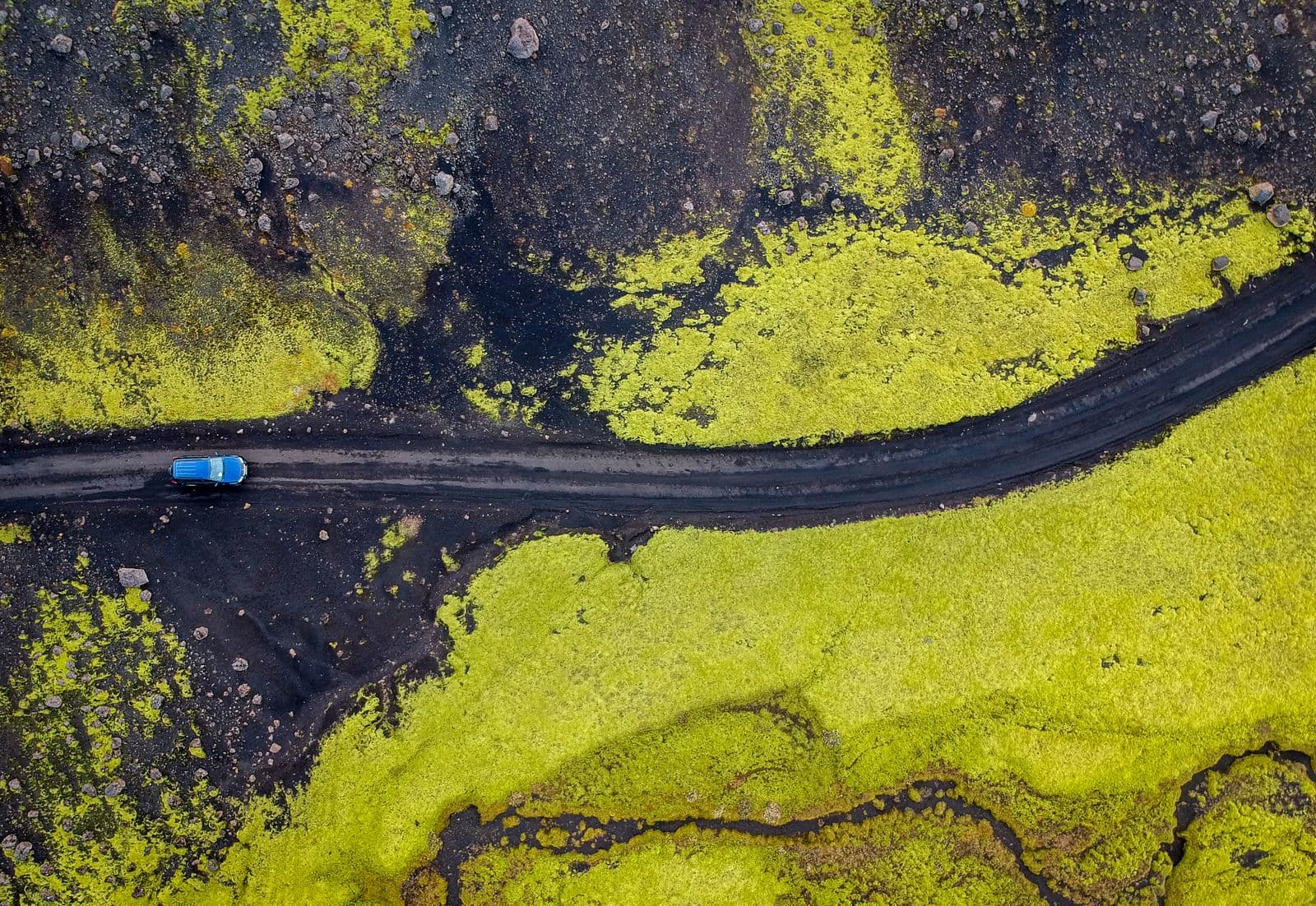 Aerial view of a car on a narrow road crossing bright green moss and black volcanic ground in Iceland
