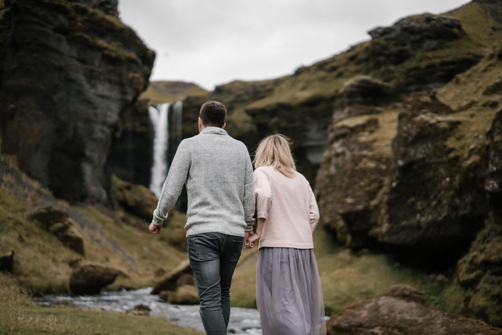 Couple holding hands walking towards a waterfall in a moss-covered canyon