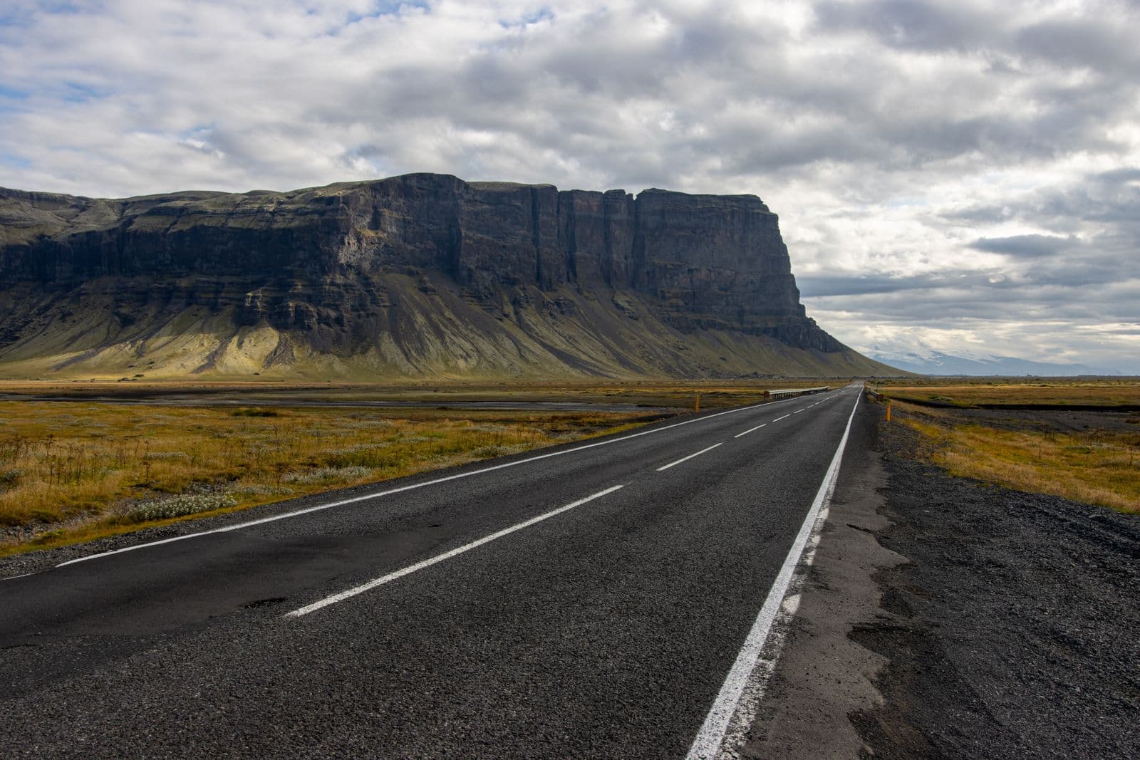 Straight road beside a huge flat-topped cliff under a cloudy sky in Iceland
