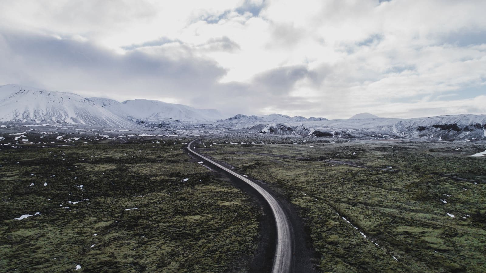 Aerial view of a winding road crossing a moss-covered lava field below snow-capped mountains