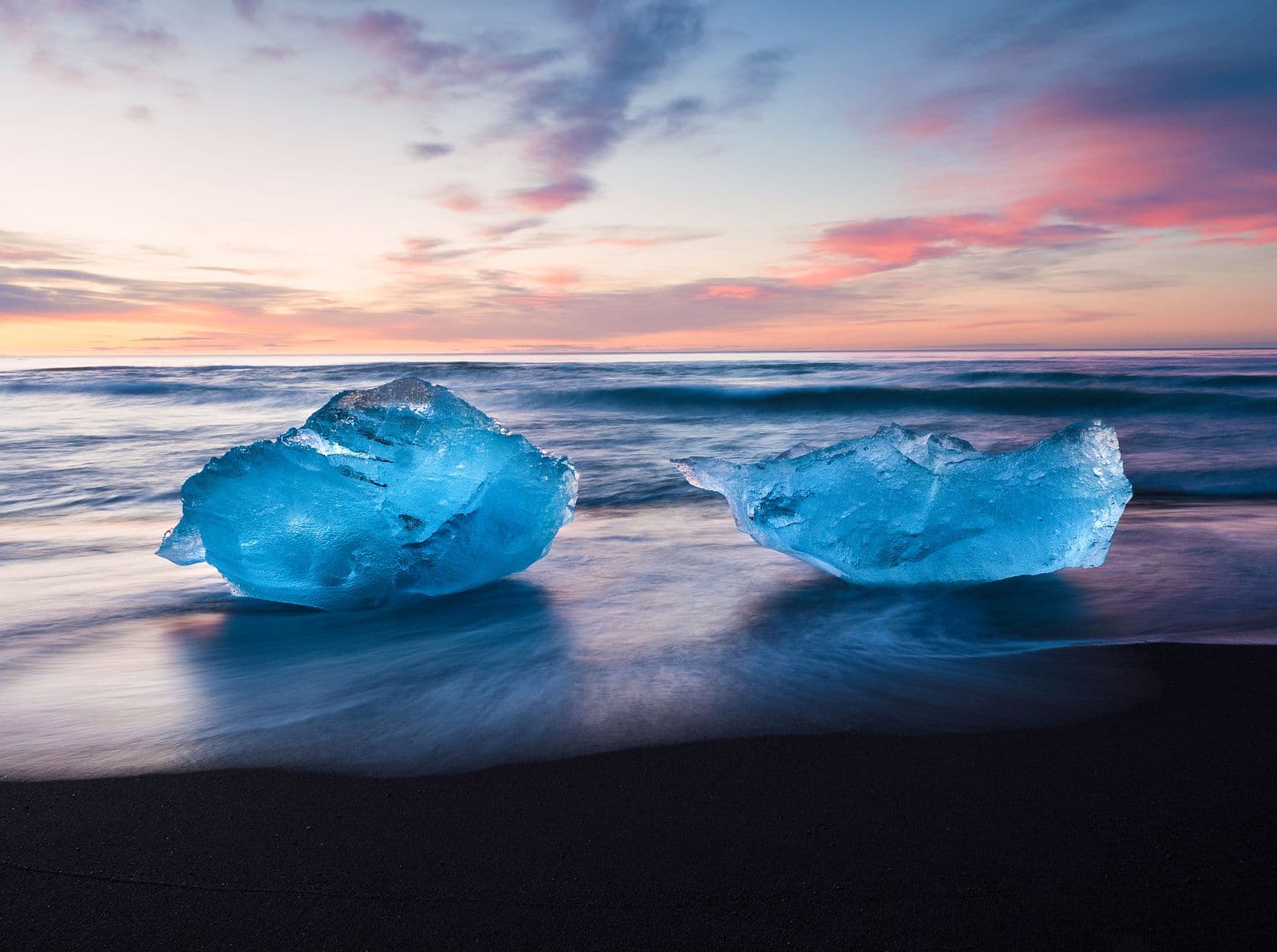Two blue pieces of ice on black sand with waves and a pink sunset sky