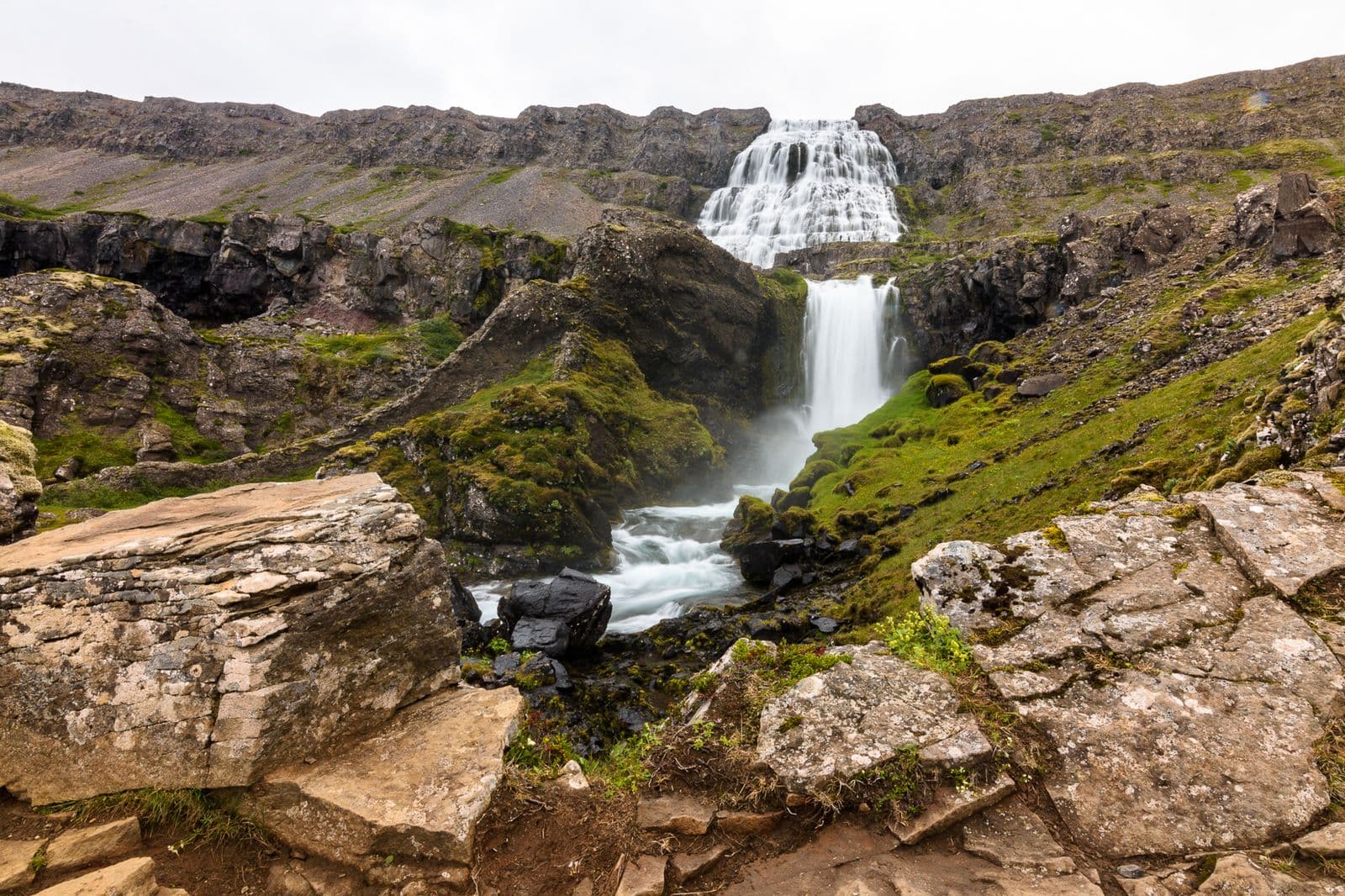 Dynjandi waterfall cascading down a wide, stepped rock face in the Westfjords, with a smaller fall and moss-covered rocks below