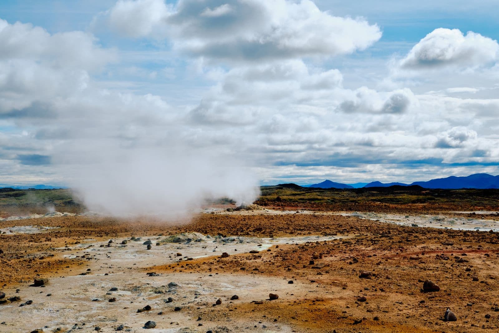 Steam rising from a geothermal field with orange ground under a partly cloudy sky