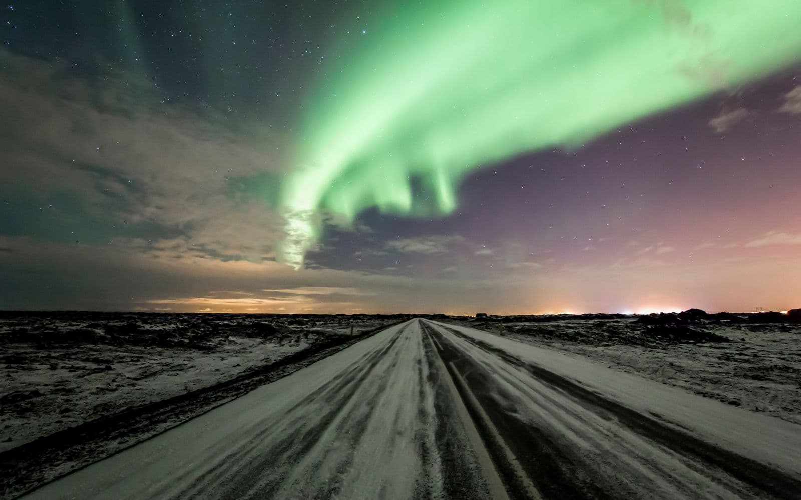 Snow-covered road under a night sky with the northern lights glowing above in Iceland