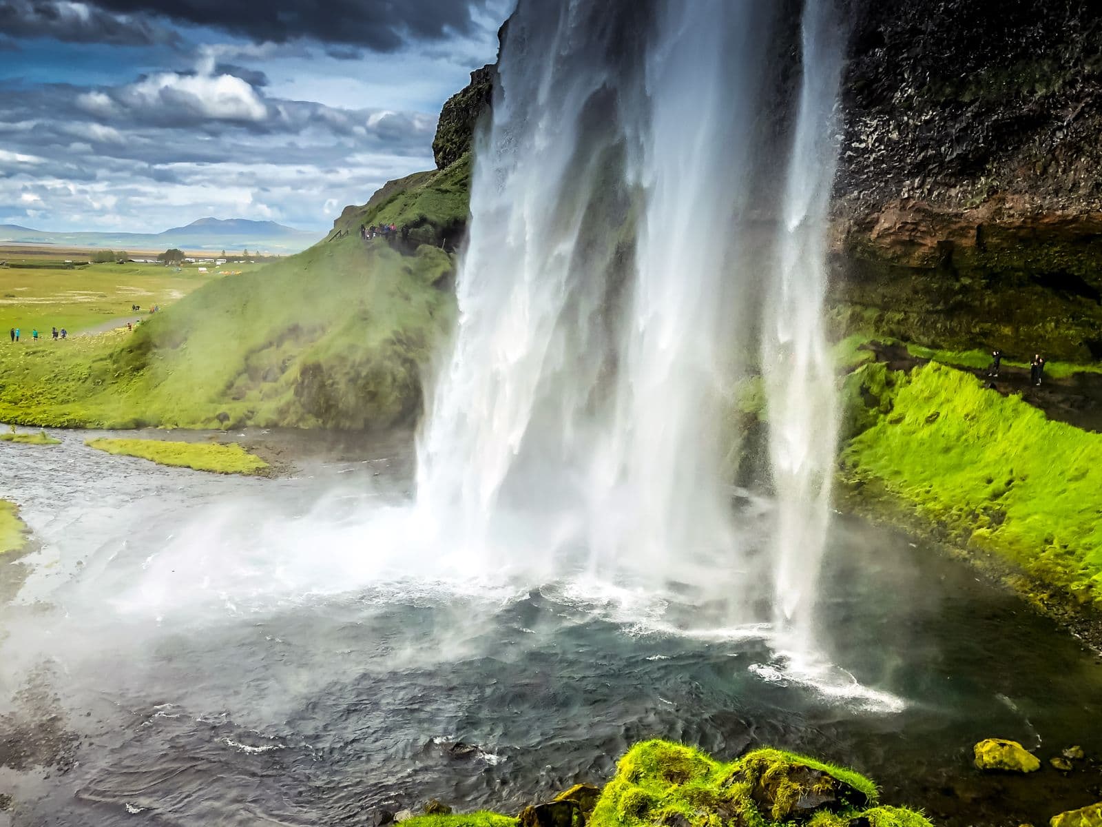 Seljalandsfoss seen from behind the falling water, with green slopes and a river below