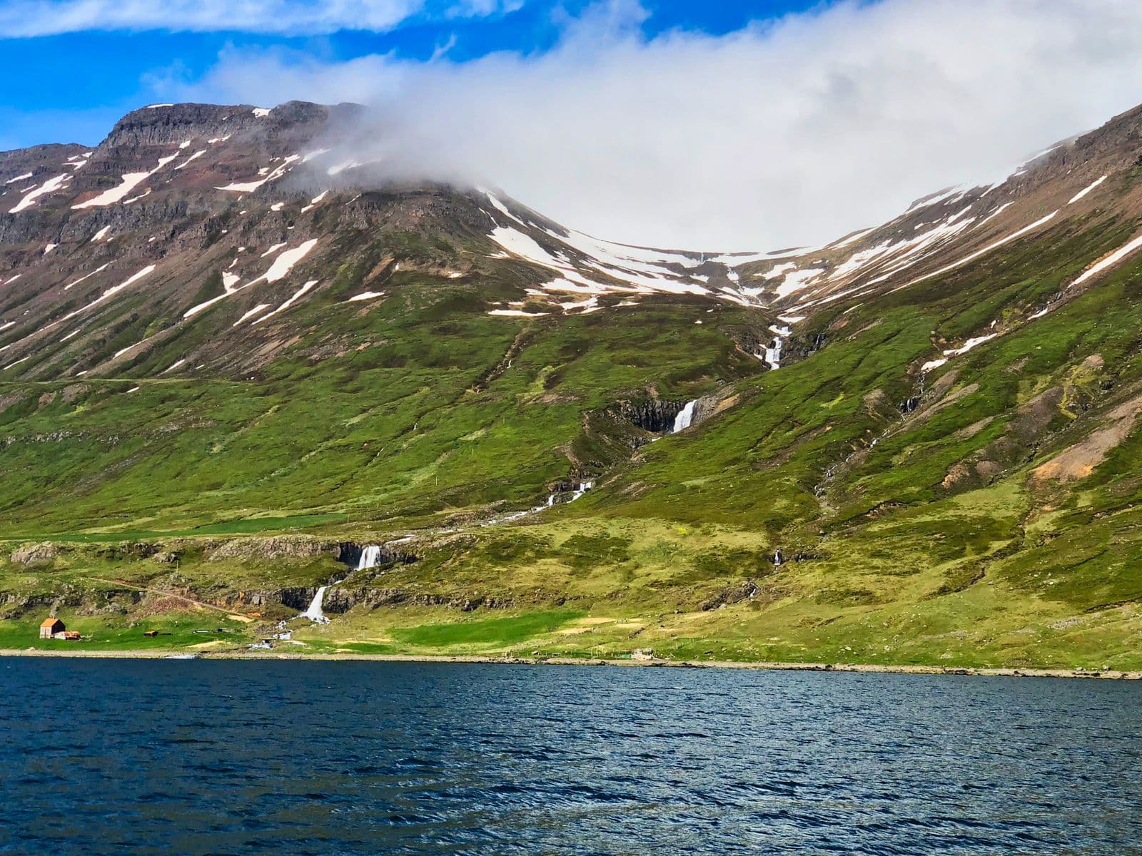 Green mountainside with snow patches and several small waterfalls above a dark blue fjord in East Iceland