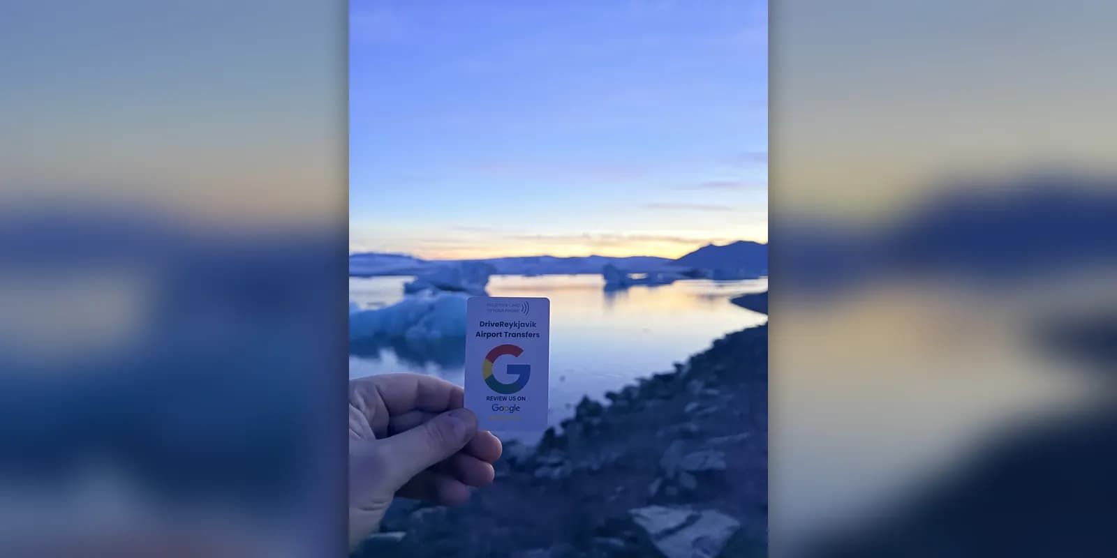 A hand holding a Drive Reykjavik review card in front of icebergs on Jökulsárlón glacier lagoon at sunset