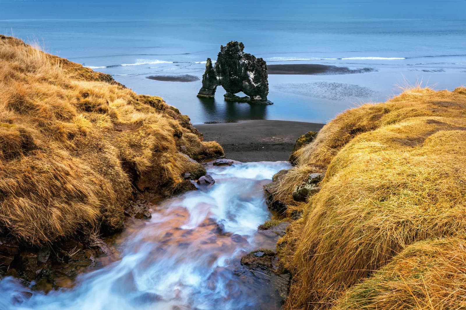 Hvítserkur sea stack seen from a grassy bank with a small stream running to the black beach